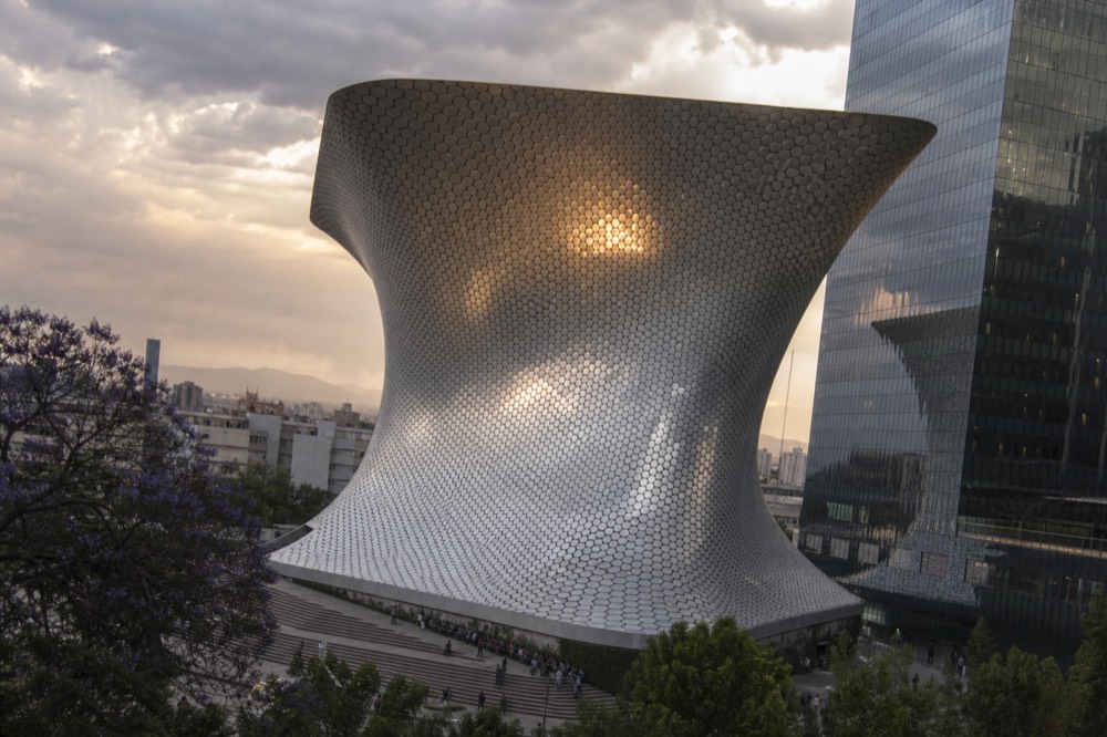 A wave-shaped mirror-clad museum building reflecting the sky in a modern city plaza